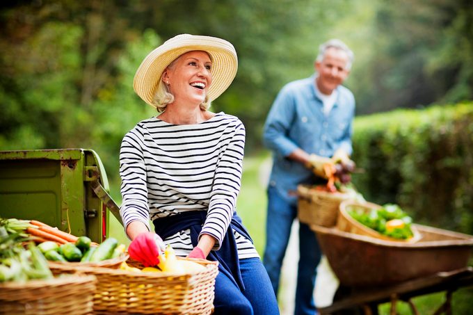 Portrait of mature woman with baskets of harvested vegetables and man working in the background. Horizontal shot.